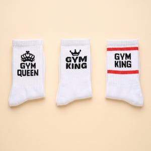 May include: Three white crew socks. The first sock features a black crown icon above the text GYM QUEEN. The second sock displays a black crown icon above the text GYM KING. The third sock has two horizontal red stripes with GYM KING text.