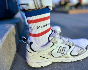 Op de afbeelding: Witte sportschoenen met zilveren accenten en witte sokken. De sokken hebben rode en blauwe strepen en de tekst "F... Monday" in het zwart. De schoenen hebben het nummer "408" aan de zijkant.