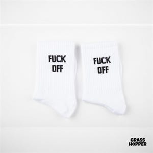 May include: A pair of white crew socks featuring the black text FUCK OFF printed on the upper portion of each sock. The socks have a ribbed texture and are displayed side by side against a plain white background with a small logo in the corner.