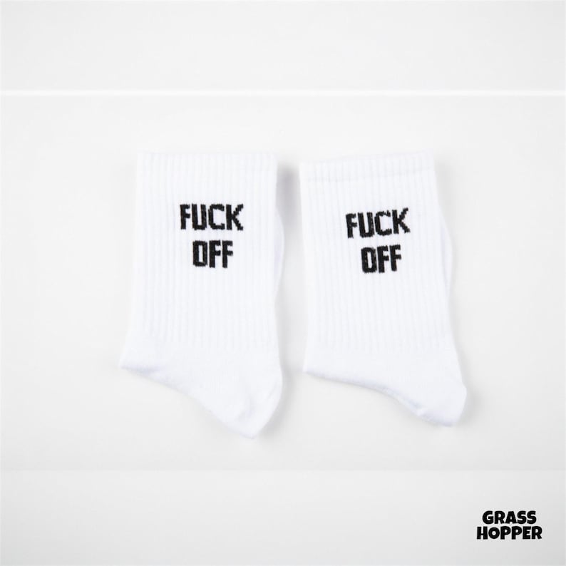 May include: A pair of white crew socks featuring the black text FUCK OFF printed on the upper portion of each sock. The socks have a ribbed texture and are displayed side by side against a plain white background with a small logo in the corner.