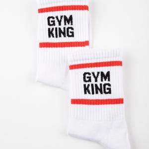 May include: A pair of white crew socks featuring two horizontal red stripes near the top cuff. The text "GYM KING" is printed in black block letters between the red stripes on each sock. The brand name "GRASS HOPPER" appears in small black text at the bottom.