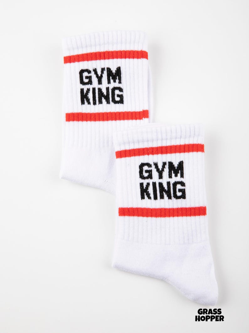 May include: A pair of white crew socks featuring two horizontal red stripes near the top cuff. The text "GYM KING" is printed in black block letters between the red stripes on each sock. The brand name "GRASS HOPPER" appears in small black text at the bottom.