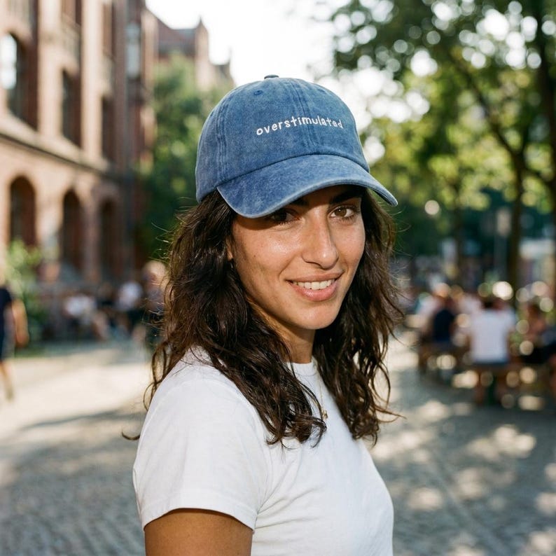 Puede incluir: Gorra de b&eacute;isbol de mezclilla azul con la palabra "overstimulated" bordada en blanco. La gorra la lleva una persona que lleva una camiseta blanca. El fondo incluye un entorno exterior borroso con &aacute;rboles y edificios.