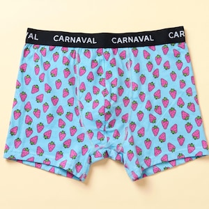 Op de afbeelding: Lichtblauwe boxershort met een herhalend patroon van roze aardbeien met groene blaadjes. De zwarte elastische tailleband toont de witte tekst CARNAVAL herhaaldelijk aan de voorzijde.