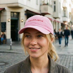 Puede incluir: Gorra de b&eacute;isbol rosa con la palabra "overstimulated" bordada en rojo. La gorra la lleva una persona con una chaqueta verde, con una escena de calle borrosa al fondo.