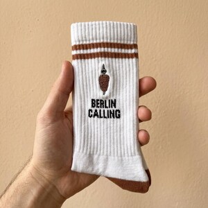 Pu&ograve; includere: Calzini bianchi a coste con dettagli marroni e la scritta "BERLIN CALLING" in nero. I calzini presentano un'immagine ricamata di un kebab marrone. Due strisce marroni sulla parte superiore del calzino.