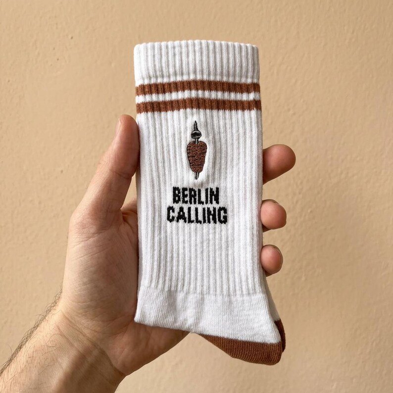Pu&ograve; includere: Calzini bianchi a coste con dettagli marroni e la scritta "BERLIN CALLING" in nero. I calzini presentano un'immagine ricamata di un kebab marrone. Due strisce marroni sulla parte superiore del calzino.