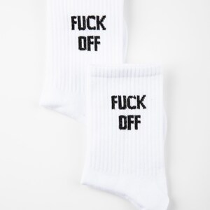 May include: Two white socks with the words "FUCK OFF" printed in black block letters. The socks are ribbed and have a casual style. The brand name "GRASSHOPPER" is printed in black at the bottom right.