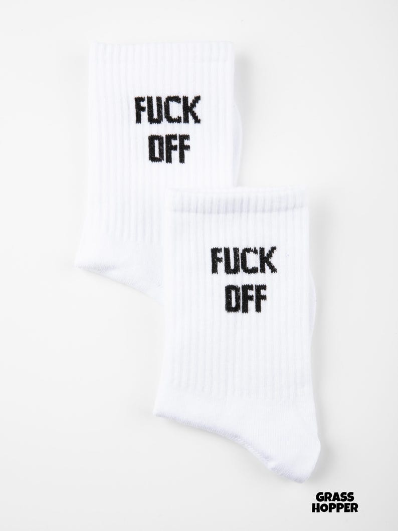 May include: Two white socks with the words "FUCK OFF" printed in black block letters. The socks are ribbed and have a casual style. The brand name "GRASSHOPPER" is printed in black at the bottom right.