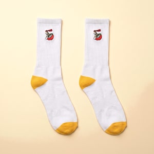 May include: A pair of white crew socks featuring a small embroidered graphic of a bowl of ramen with red, green, and yellow details. The socks have a ribbed cuff, a solid yellow heel, and a solid yellow toe section, providing a distinct colour contrast.
