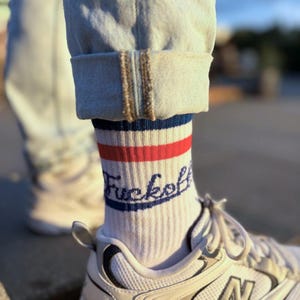 Könnte beinhalten: Weiße Socken mit roten und blauen Streifen und dem Wort "Fuckoff" in marineblauer Schrift. Die Socken werden mit hellblauen Jeans und weißen Turnschuhen mit silbernen Akzenten getragen. Der Markenname "Grass Hopper" ist sichtbar.