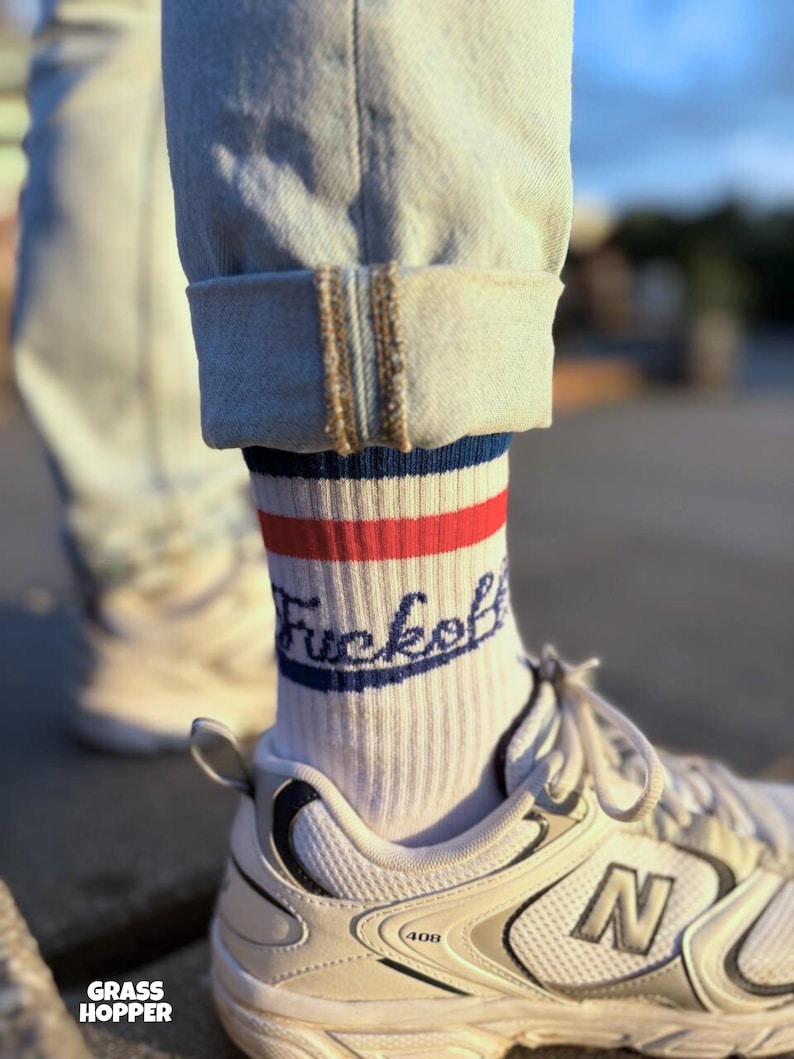 May include: White socks with red and blue stripes and the word "Fuckoff" in navy blue script. The socks are worn with light-wash denim jeans and white sneakers with silver accents. The brand name "Grass Hopper" is visible.