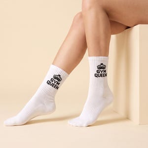 May include: A pair of white crew socks featuring a black graphic print on the upper cuff. The design includes a stylized crown icon positioned above the text "GYM QUEEN" in a bold, sans-serif font. The socks have a ribbed texture.
