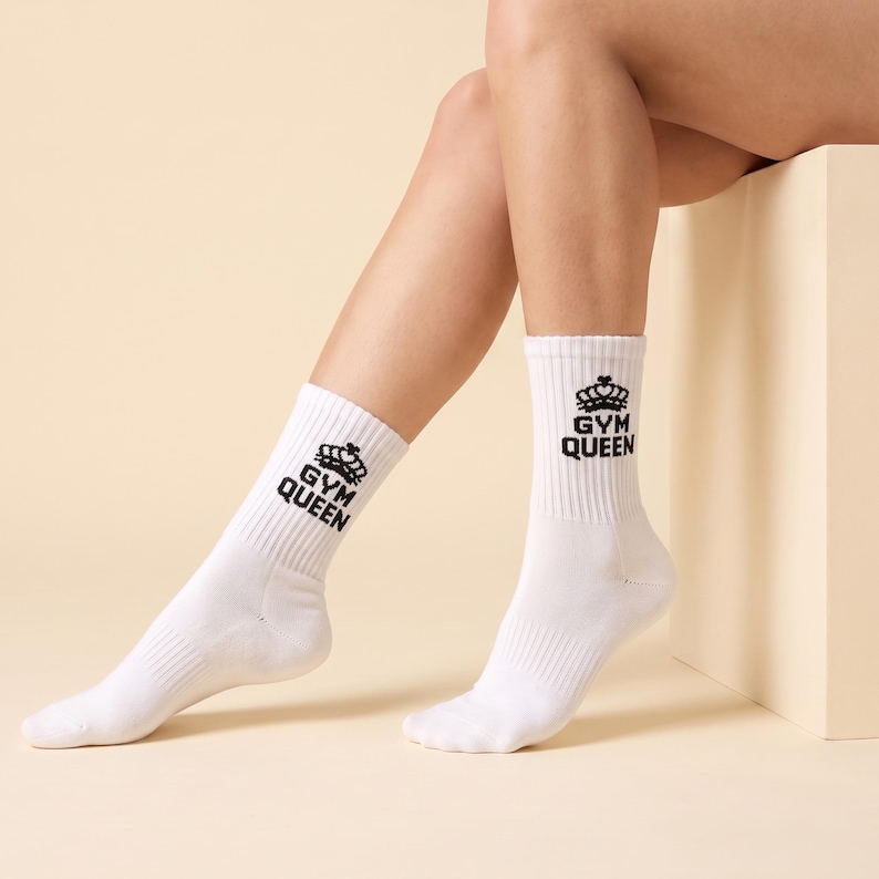 May include: A pair of white crew socks featuring a black graphic print on the upper cuff. The design includes a stylized crown icon positioned above the text "GYM QUEEN" in a bold, sans-serif font. The socks have a ribbed texture.