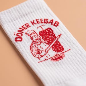 Pu&ograve; includere: Calzini bianchi a coste con grafica rossa "D&ouml;ner Kebab" raffigurante uno chef e uno spiedo per kebab. I calzini sono realizzati in un materiale morbido e confortevole.