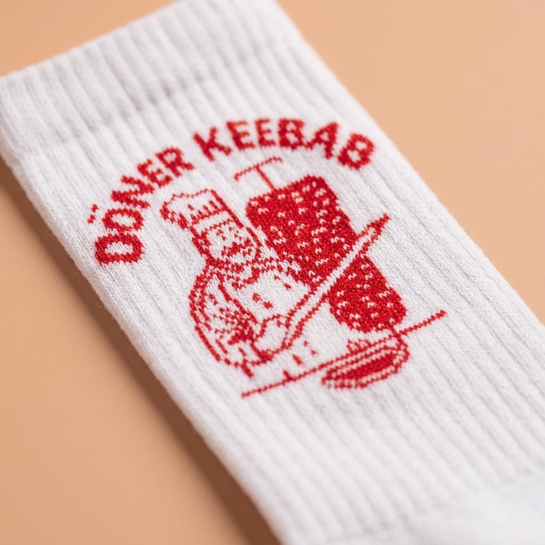 Pu&ograve; includere: Calzini bianchi a coste con grafica rossa "D&ouml;ner Kebab" raffigurante uno chef e uno spiedo per kebab. I calzini sono realizzati in un materiale morbido e confortevole.