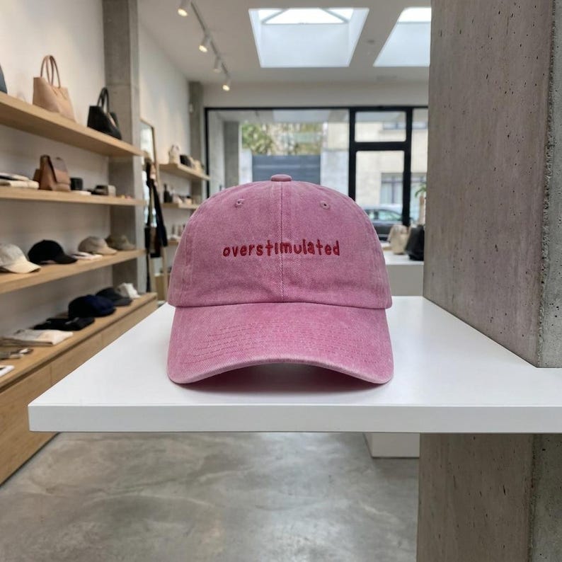 Puede incluir: Gorra de b&eacute;isbol rosa con la palabra "overstimulated" bordada en rojo en la parte delantera. La gorra est&aacute; hecha de una tela suave y texturizada y tiene una visera curva. El sombrero se exhibe en un estante blanco.