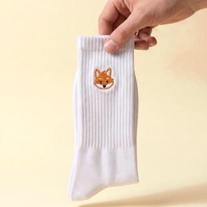 Könnte beinhalten: Eine weiße, gerippte Socke mit einem kleinen, aufgestickten orange-weißen Fuchskopf in der Mitte des oberen Bereichs. Das Design umfasst detaillierte Stickereien für die Ohren, Augen und Schnauze des Fuchses auf dem schlichten weißen Stoff.