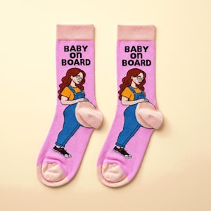 May include: A pair of light pink crew socks featuring a graphic of a person with long wavy brown hair, glasses, a yellow shirt, and blue overalls. The text BABY ON BOARD is printed in black above the figure, with light pink cuffs and heels.