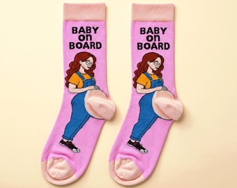 May include: A pair of light pink crew socks featuring a graphic of a person with long wavy brown hair, glasses, a yellow shirt, and blue overalls. The text BABY ON BOARD is printed in black above the figure, with light pink cuffs and heels.