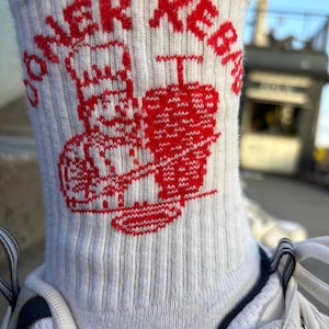 Pu&ograve; includere: Calzini bianchi a coste con una grafica rossa di uno chef e un kebab, insieme al testo "DONER KEBAB". I calzini sono indossati con scarpe da ginnastica bianche. La parola "GRASSHOPPER" &egrave; stampata in basso.
