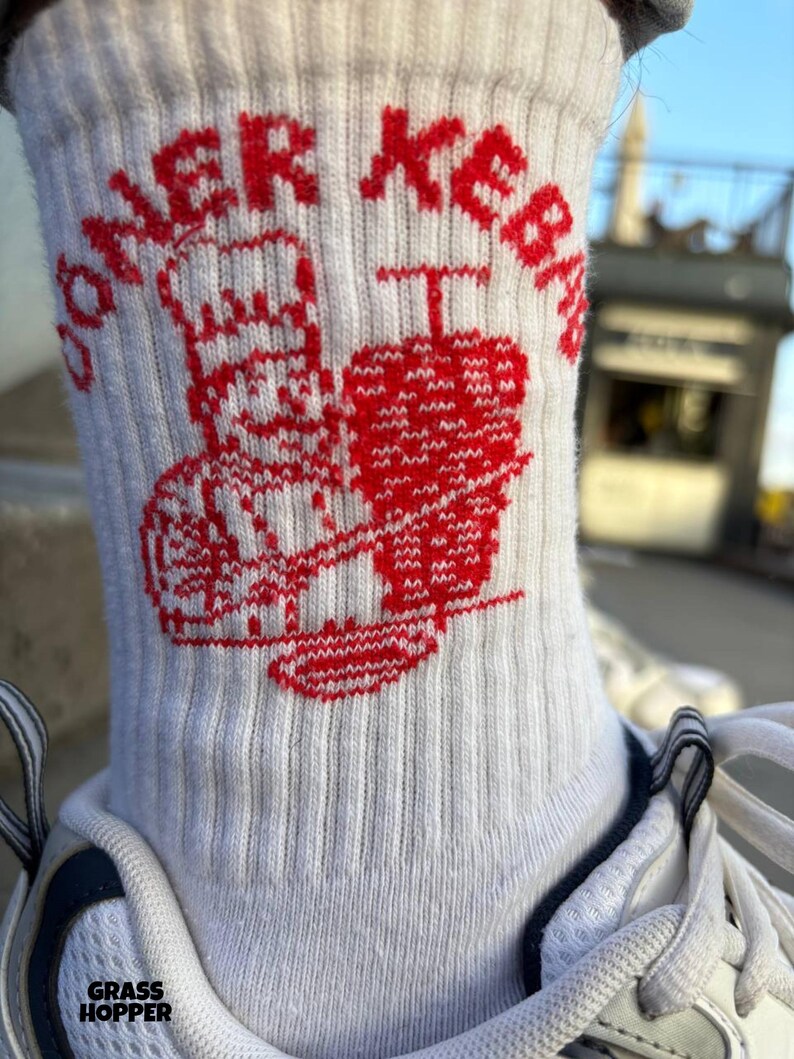 Pu&ograve; includere: Calzini bianchi a coste con una grafica rossa di uno chef e un kebab, insieme al testo "DONER KEBAB". I calzini sono indossati con scarpe da ginnastica bianche. La parola "GRASSHOPPER" &egrave; stampata in basso.