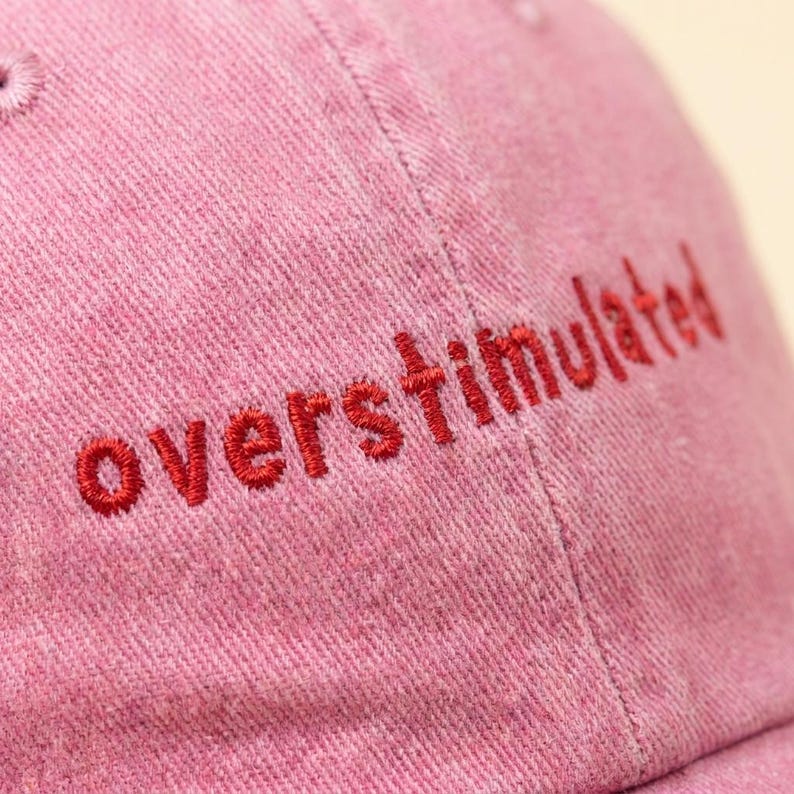 Puede incluir: Gorra de b&eacute;isbol rosa polvoriento con la palabra "overstimulated" bordada en hilo rojo. La gorra est&aacute; hecha de una tela texturizada y tiene una visera curva.