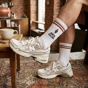 Pu&ograve; includere: Sneakers New Balance color crema con design a rete e la lettera "N" sul lato. Le sneakers sono abbinate a calzini bianchi con strisce marroni e la scritta "Berlin Calling". Una tazza di caff&egrave; &egrave; appoggiata su un tavolo di legno.