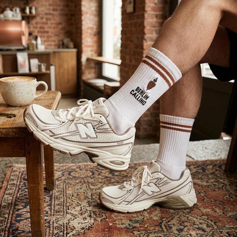 Pu&ograve; includere: Sneakers New Balance color crema con design a rete e la lettera "N" sul lato. Le sneakers sono abbinate a calzini bianchi con strisce marroni e la scritta "Berlin Calling". Una tazza di caff&egrave; &egrave; appoggiata su un tavolo di legno.