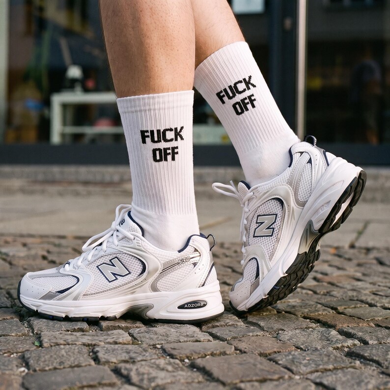 May include: White crew socks featuring the black text FUCK OFF in a bold, blocky font, paired with white, silver, and navy blue New Balance 530 athletic sneakers with mesh panels, white laces, and visible ADZORB branding on the soles.