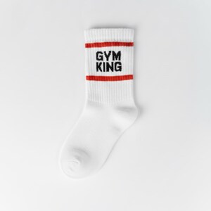 Pode incluir: Meia atl&eacute;tica branca com listras vermelhas e o texto "GYM KING" em preto. A meia &eacute; feita de um material canelado e foi projetada para conforto e desempenho. A meia &eacute; de comprimento de tripula&ccedil;&atilde;o.