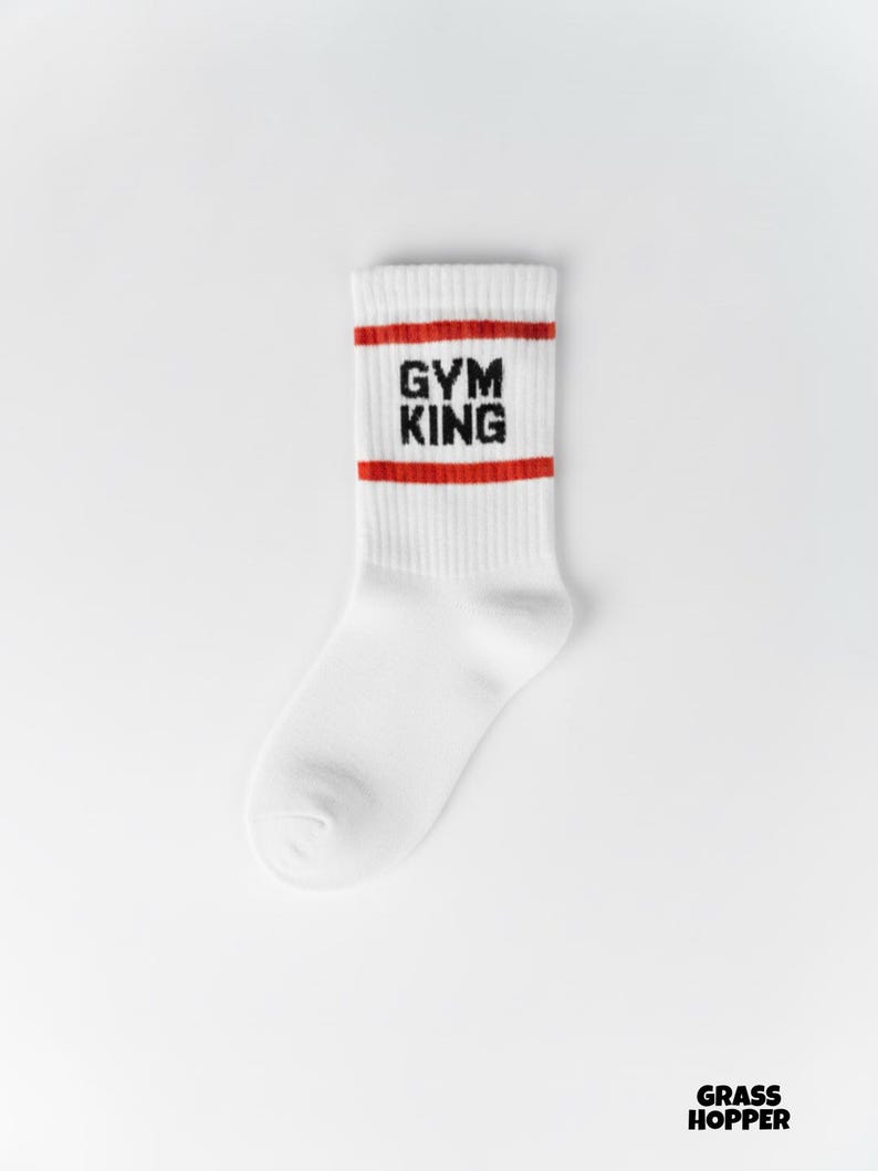 Pode incluir: Meia atl&eacute;tica branca com listras vermelhas e o texto "GYM KING" em preto. A meia &eacute; feita de um material canelado e foi projetada para conforto e desempenho. A meia &eacute; de comprimento de tripula&ccedil;&atilde;o.