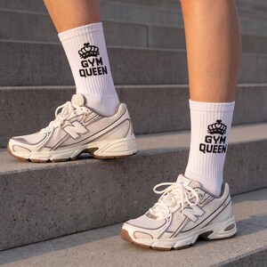 May include: A pair of white crew socks featuring a black crown graphic and the text GYM QUEEN in a bold font, worn with white and beige athletic sneakers that have a mesh upper, white laces, and a brown rubber outsole on the bottom.