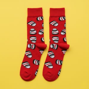 Puede incluir: Un par de calcetines rojos con un patrón repetido de recipientes blancos y negros, cada uno con la etiqueta "Yummy". Los calcetines tienen un puño y una puntera rojos lisos. El fondo es amarillo liso.