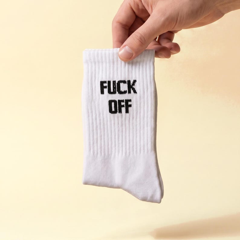 May include: A white ribbed crew sock featuring the text FUCK OFF printed in bold black capital letters on the front. The sock is made of a knit fabric and has a standard length that extends above the ankle with a reinforced cuff at the top.