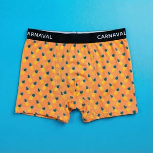 Może przedstawiać: Pomarańczowe bokserki z żółtym wzorem ananasa i niebieskimi akcentami. Pas jest czarny z napisem "ARNAVAL" z przodu i "CARNAVAL" z tyłu. Szorty wykonane są z miękkiego materiału.