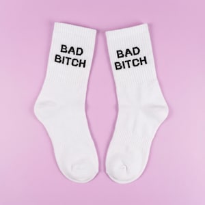 Könnte beinhalten: Ein Paar weiße Socken mit dem schwarzen Aufdruck "BAD BITCH". Die Socken sind auf einem hellvioletten Hintergrund platziert. Die Socken bestehen aus einem weichen, gerippten Material.