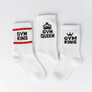 Pode incluir: Tr&ecirc;s pares de meias esportivas brancas com texto e gr&aacute;ficos pretos. Um par tem "GYM KING" em preto com uma faixa vermelha. Outro par diz "GYM QUEEN" com um gr&aacute;fico de coroa. O terceiro par diz "GYM KING" com uma coroa.