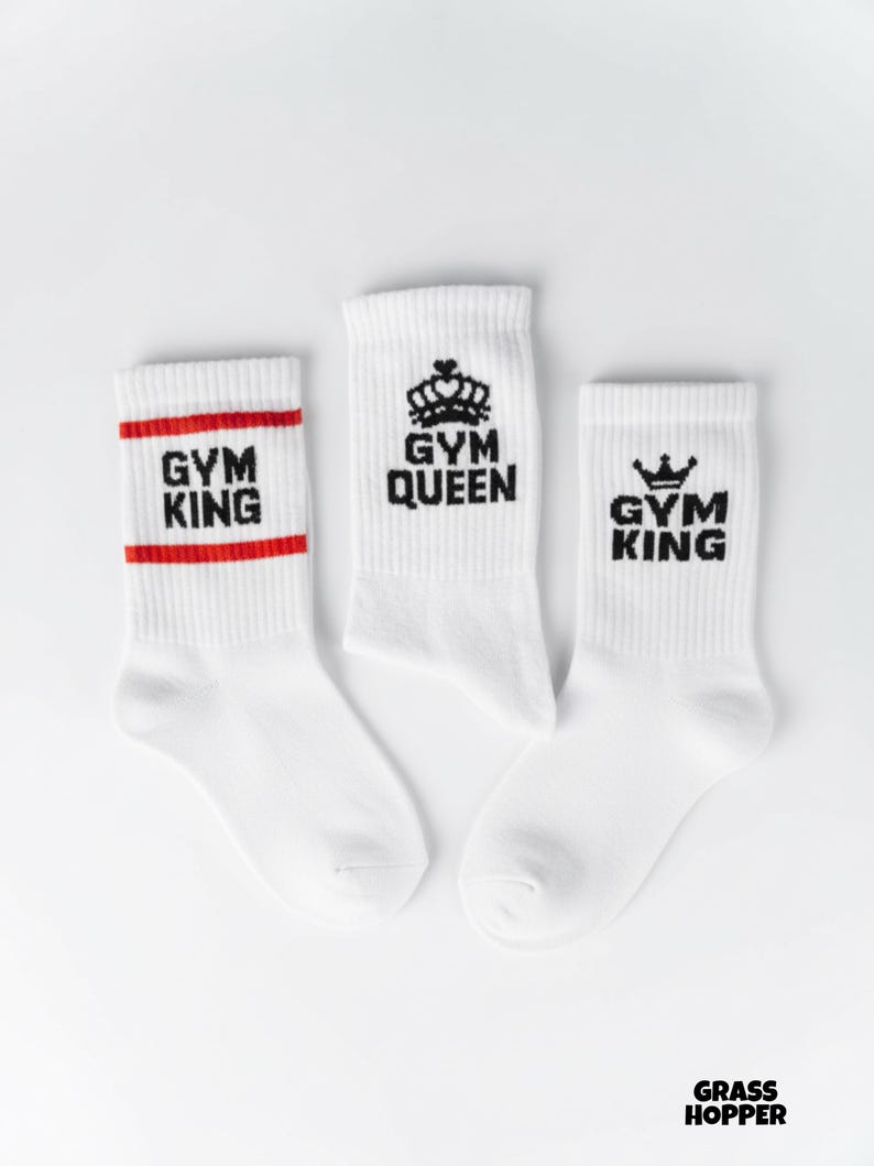 Pode incluir: Tr&ecirc;s pares de meias esportivas brancas com texto e gr&aacute;ficos pretos. Um par tem "GYM KING" em preto com uma faixa vermelha. Outro par diz "GYM QUEEN" com um gr&aacute;fico de coroa. O terceiro par diz "GYM KING" com uma coroa.