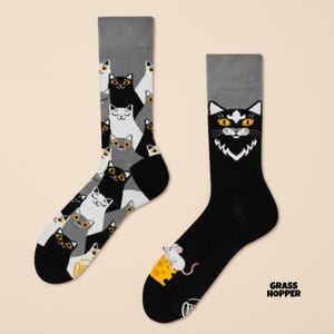 Könnte beinhalten: Ein Paar Socken mit Katzenmotiv. Eine Socke zeigt ein Muster aus schwarzen, weißen und grauen Katzen. Die andere Socke ist schwarz mit einem Katzenkopf und einer Maus mit Käse. Die Socken haben graue Bündchen. Der Text "GRASS HOPPER" befindet sich unten rechts.