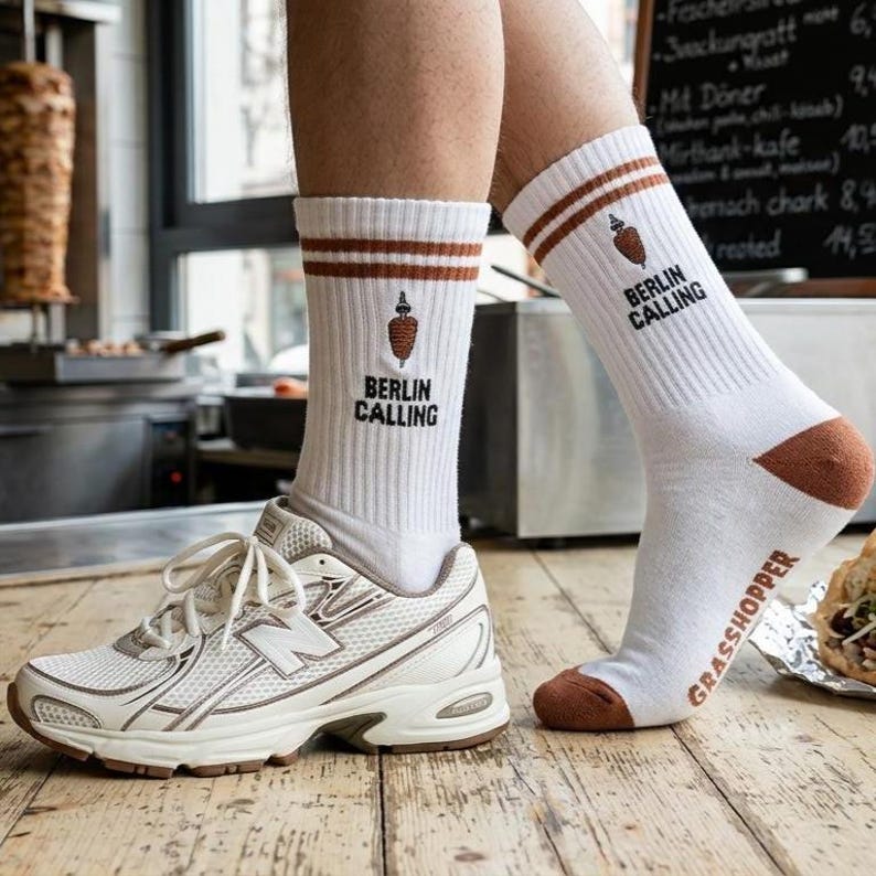 Pu&ograve; includere: Calzini bianchi con dettagli marroni, con la scritta "BERLIN CALLING" e una grafica di kebab. I calzini hanno strisce marroni nella parte superiore e talloni e punte marroni. La parola "GRASSHOPPER" &egrave; stampata sul lato.