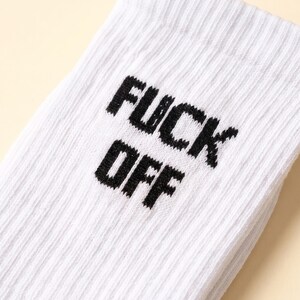 May include: A white ribbed crew sock featuring the text FUCK OFF printed in bold black capital letters. The fabric has a distinct vertical texture, and the lettering is positioned clearly on the upper portion of the sock.