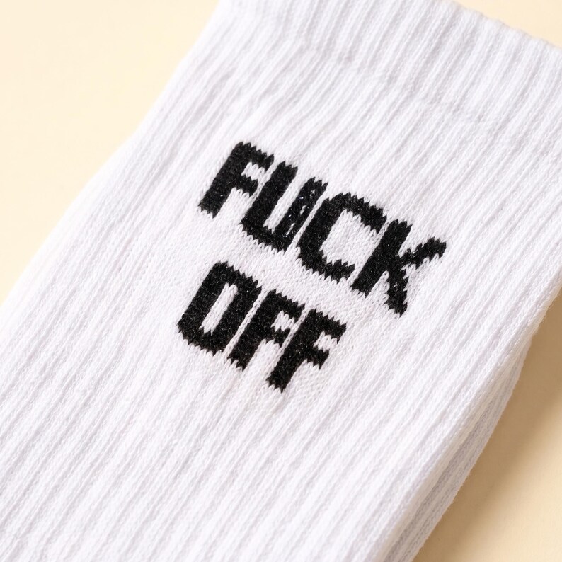 May include: A white ribbed crew sock featuring the text FUCK OFF printed in bold black capital letters. The fabric has a distinct vertical texture, and the lettering is positioned clearly on the upper portion of the sock.