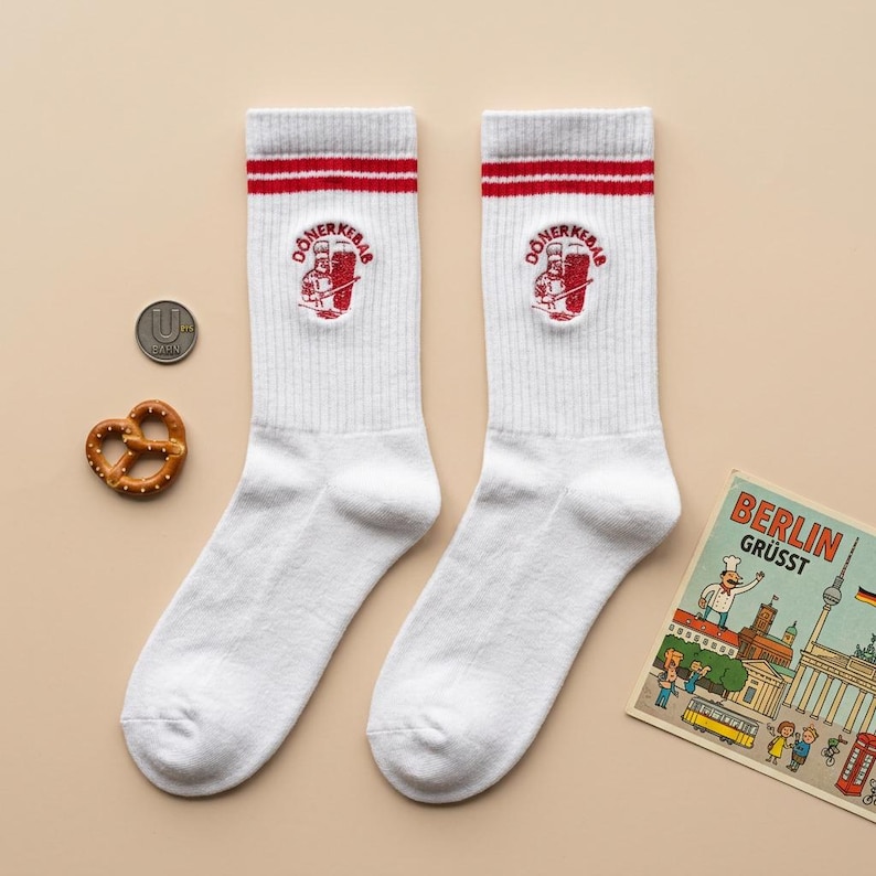 Pu&ograve; includere: Calzini bianchi con strisce rosse e una toppa "D&ouml;ner Kebab". Un pretzel e una moneta sono accanto ai calzini. Una cartolina con la scritta "BERLIN GR&Uuml;SST" &egrave; presente nell'immagine.