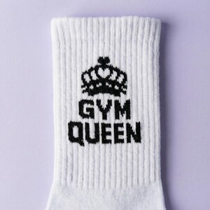 May include: A white ribbed crew sock featuring a black graphic of a crown positioned above the text GYM QUEEN. The design is centered on the front of the sock, which has a vertical ribbed texture throughout the fabric construction.