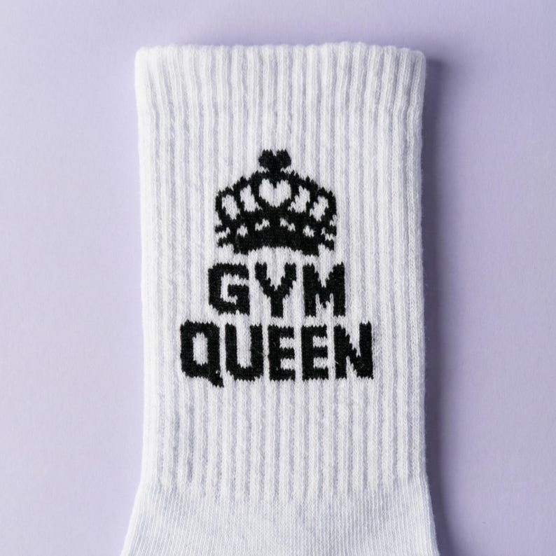 May include: A white ribbed crew sock featuring a black graphic of a crown positioned above the text GYM QUEEN. The design is centered on the front of the sock, which has a vertical ribbed texture throughout the fabric construction.
