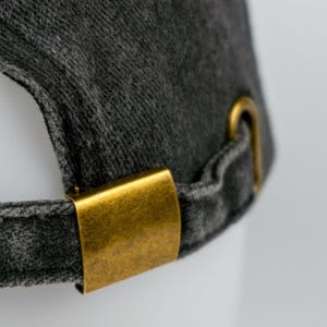 May include: Close-up of a dark gray baseball cap with a curved brim. The cap features a brass-colored metal clasp and adjustment hardware. The fabric has a textured appearance. The text "GRASS HOPPER" is visible in the lower right corner.