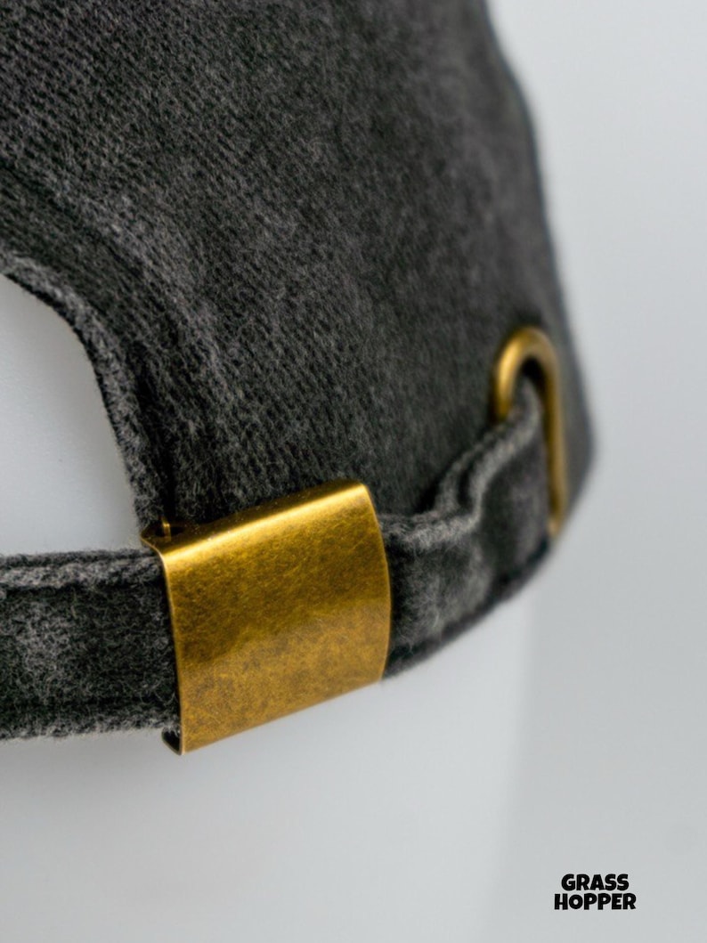 May include: Close-up of a dark gray baseball cap with a curved brim. The cap features a brass-colored metal clasp and adjustment hardware. The fabric has a textured appearance. The text "GRASS HOPPER" is visible in the lower right corner.