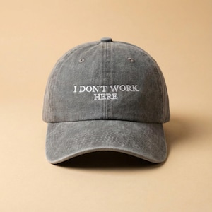 Op de afbeelding: Een grijze baseballcap met de geborduurde tekst "I DON'T WORK HERE" in het wit. De pet heeft een gebogen klep en een knoop aan de bovenkant. De hoed is gemaakt van een zacht, versleten materiaal.
