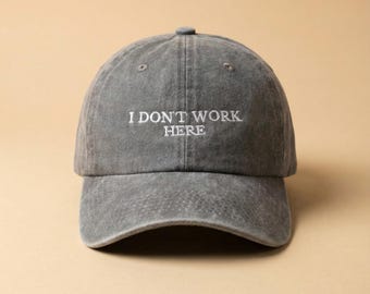 Op de afbeelding: Een grijze baseballcap met de geborduurde tekst "I DON'T WORK HERE" in het wit. De pet heeft een gebogen klep en een knoop aan de bovenkant. De hoed is gemaakt van een zacht, versleten materiaal.